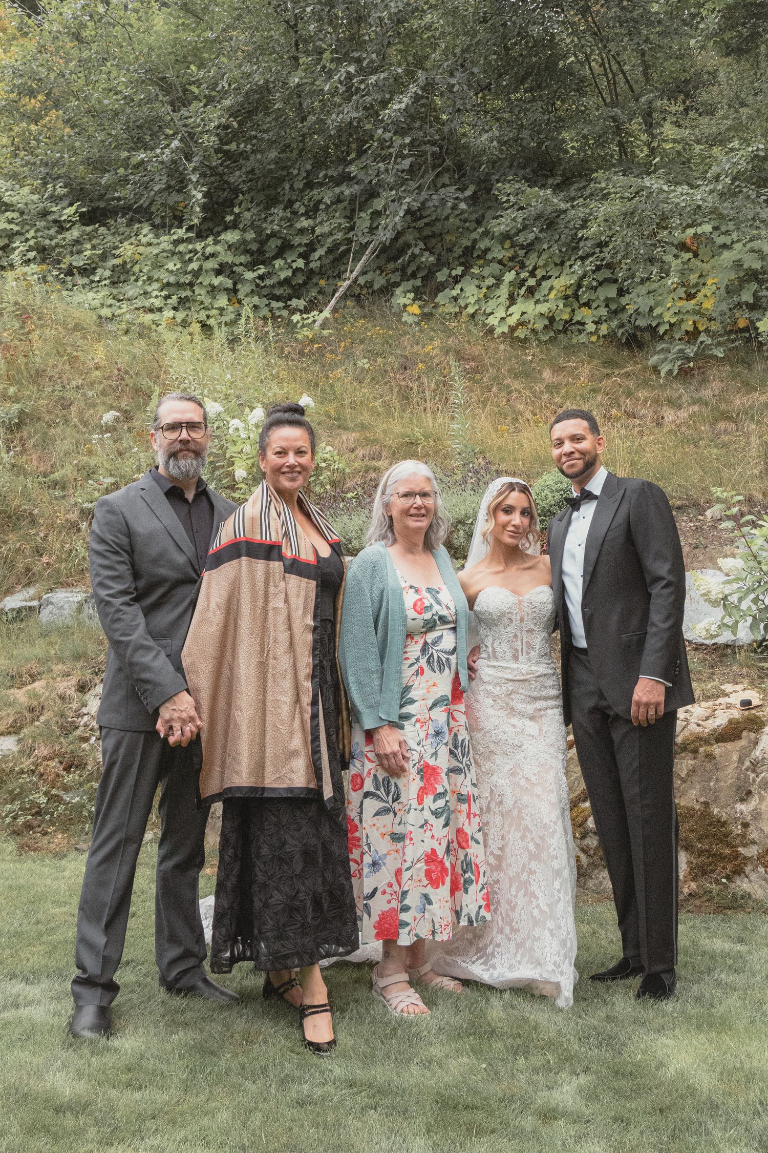 Wedding group photo with bride in lace gown, groom in tuxedo, and three guests posing outdoors on green lawn.