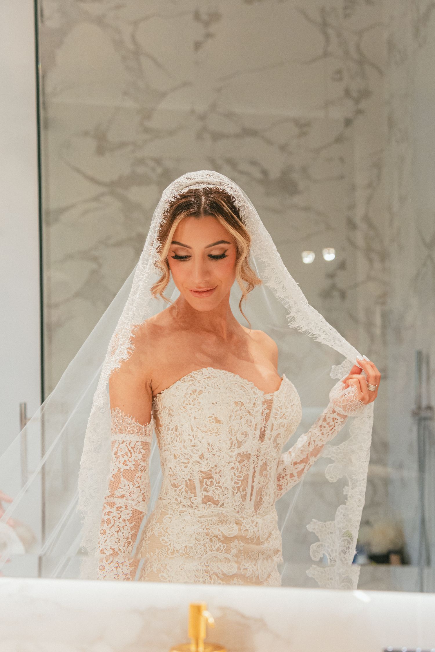 Bride in lace strapless gown with long sleeves and cathedral veil, posing elegantly in a marble bathroom.