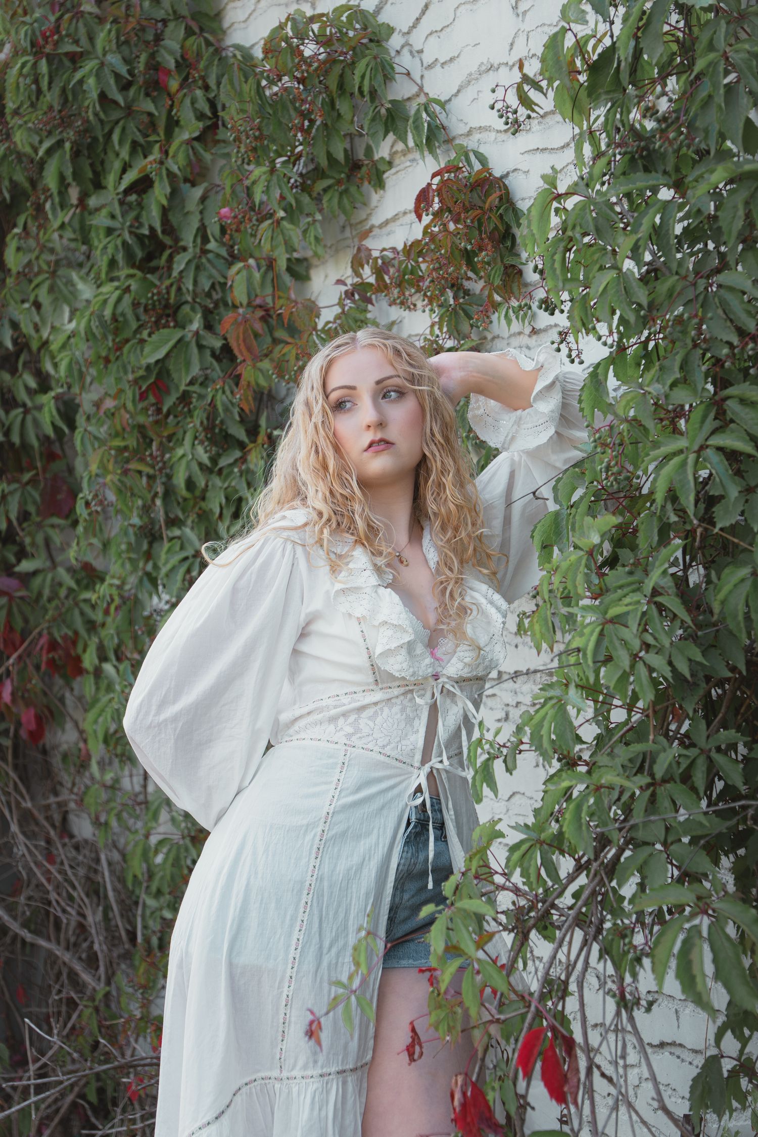 Blonde curly-haired woman in white boho dress and denim shorts posing among lush green and red ivy vines.