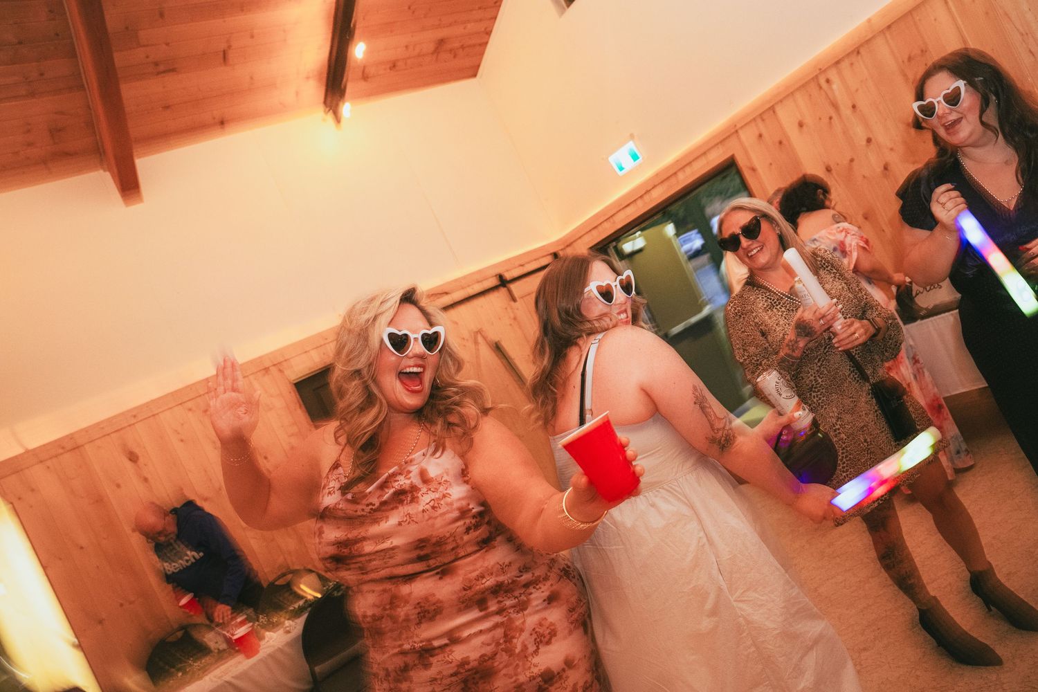 Women in sunglasses celebrating at a wedding reception party, holding glow sticks and cups in a wood-paneled venue.