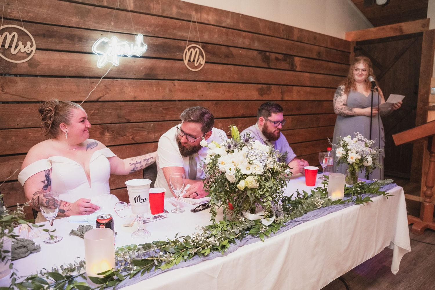 Bride and groom sit at the head table during wedding reception in a rustic barn venue with floral decor.