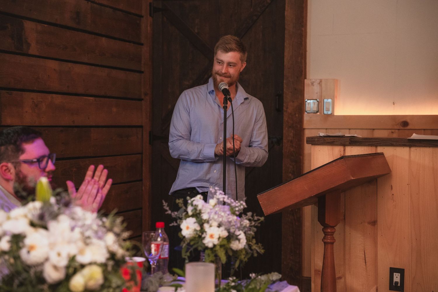 Man delivers a heartfelt wedding speech at a podium in a rustic barn reception venue with floral arrangements.