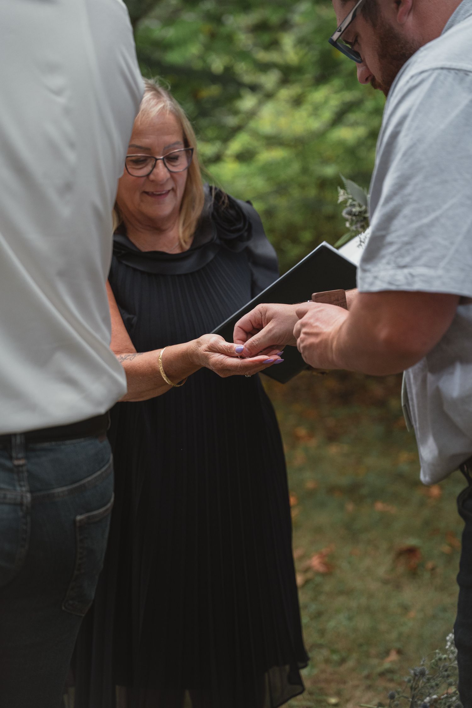 Smiling officiant in black dress oversees ring exchange between couple at outdoor wedding.