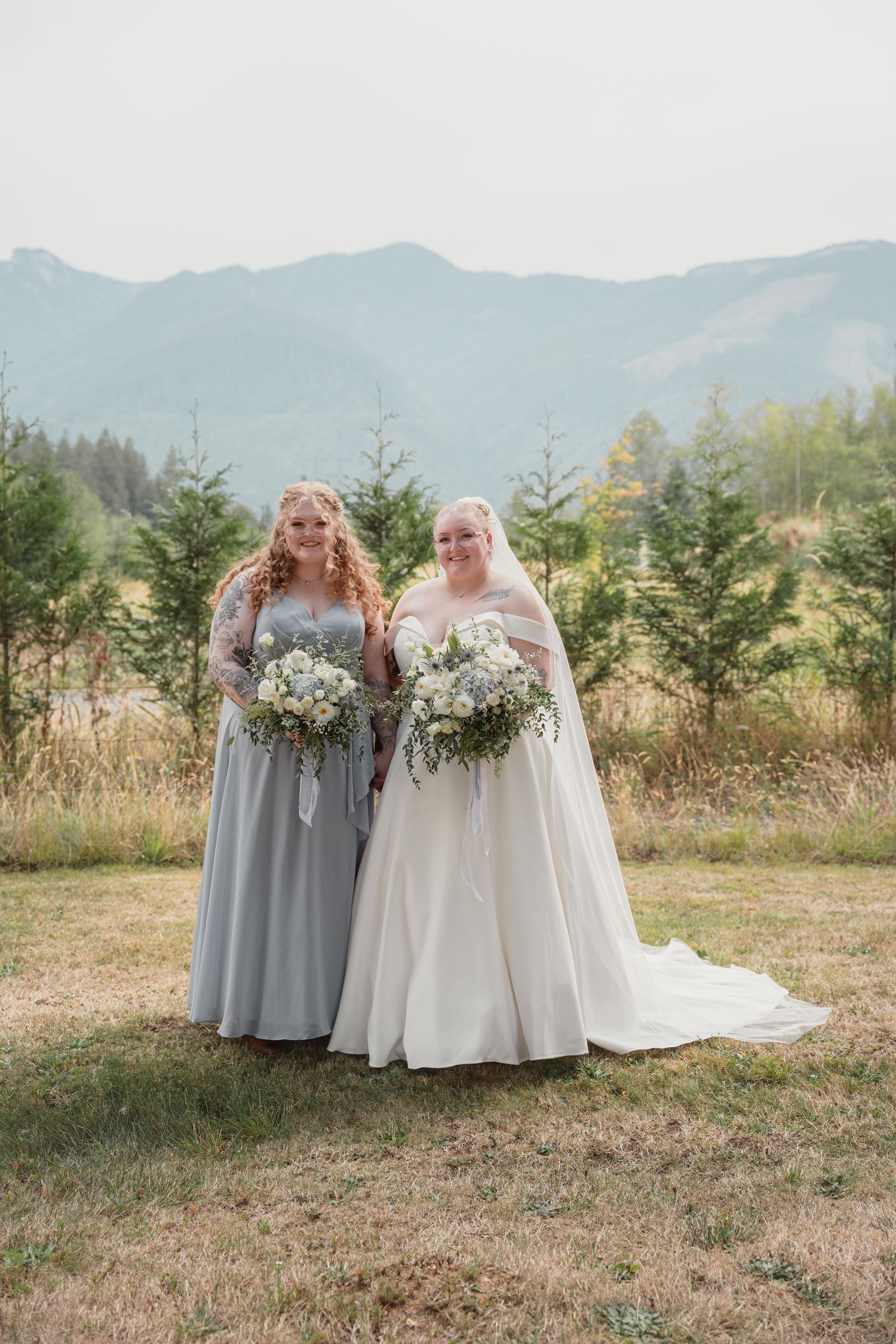 Bride in white gown and bridesmaid in gray dress holding bouquets with mountain backdrop.