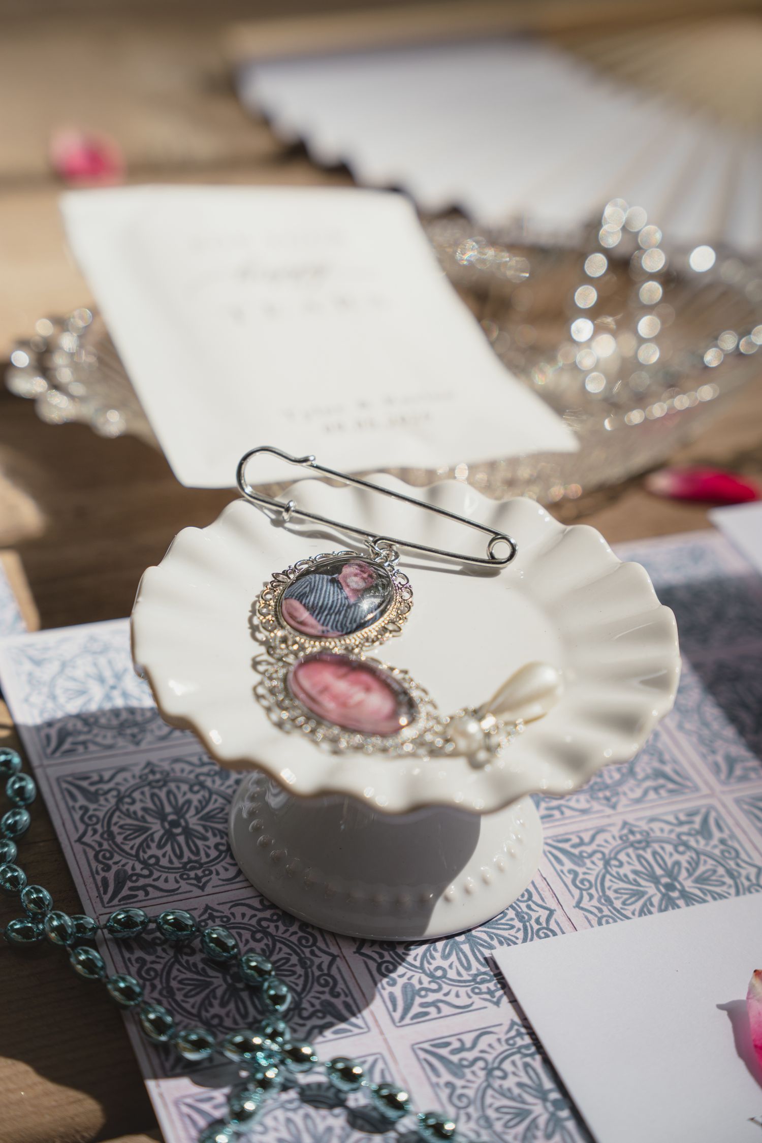 White ceramic cake stand holding vintage photo brooches and safety pin, surrounded by wedding invitations, tiara, and beaded necklace.