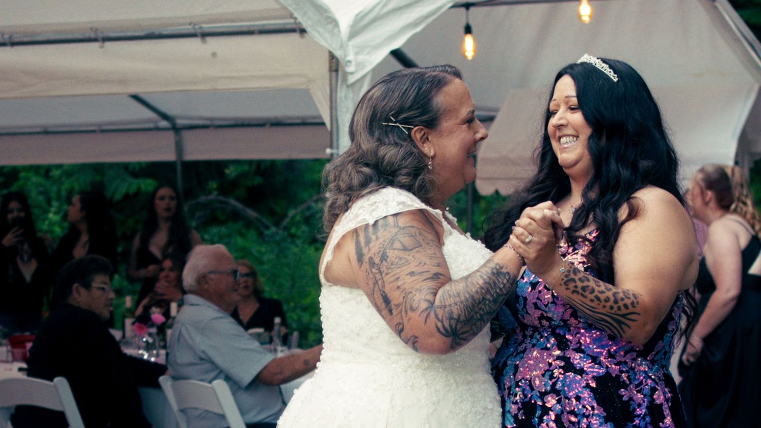 Two women share a joyful dance together at an outdoor wedding reception under a white tent.