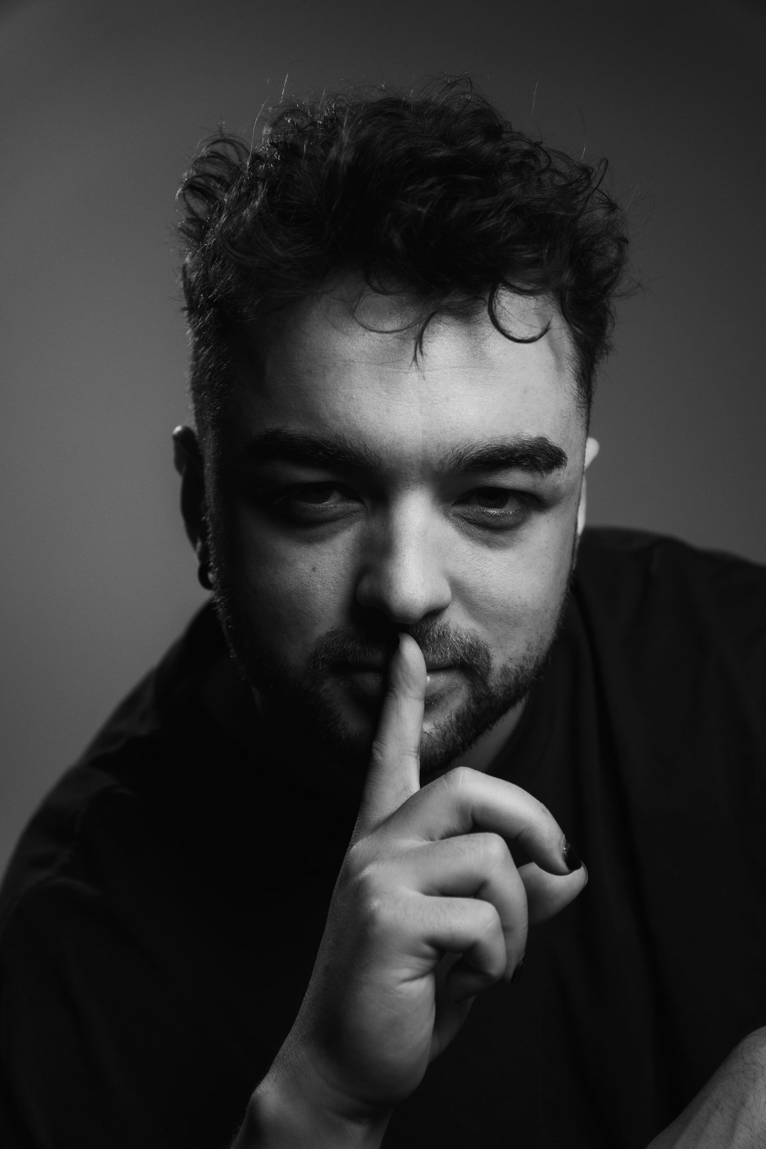 Black and white portrait of bearded man making silence gesture with finger to lips, dramatic studio lighting.
