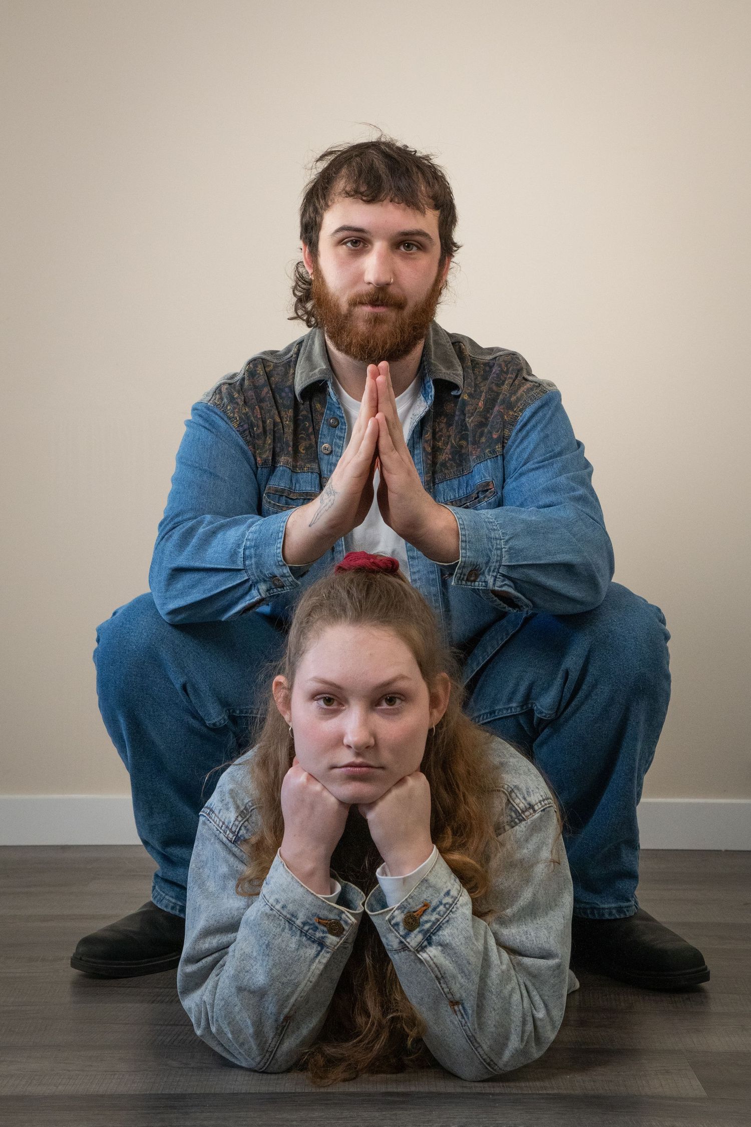 Man and woman in matching denim outfits posing indoors, man squatting with hands together, woman lying in front.