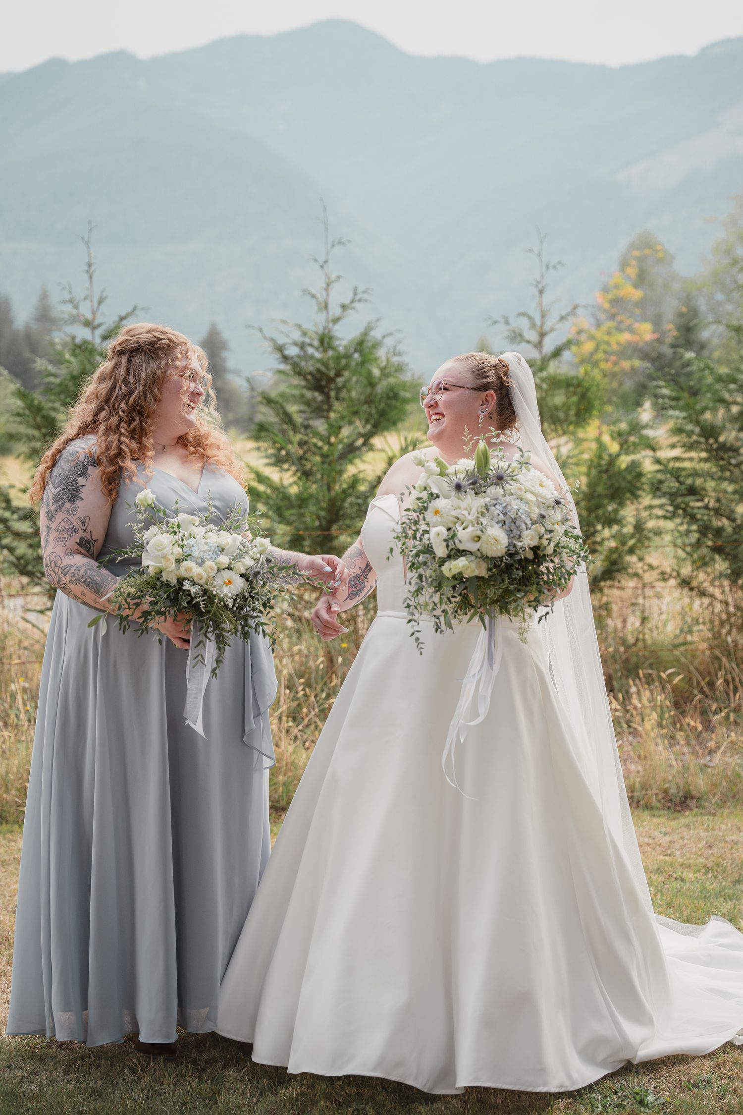 Smiling bride and bridesmaid holding hands with bouquets in mountain setting.