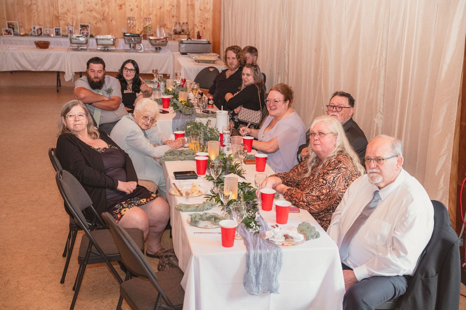Wedding guests smile at camera while seated at decorated reception table with red cups.
