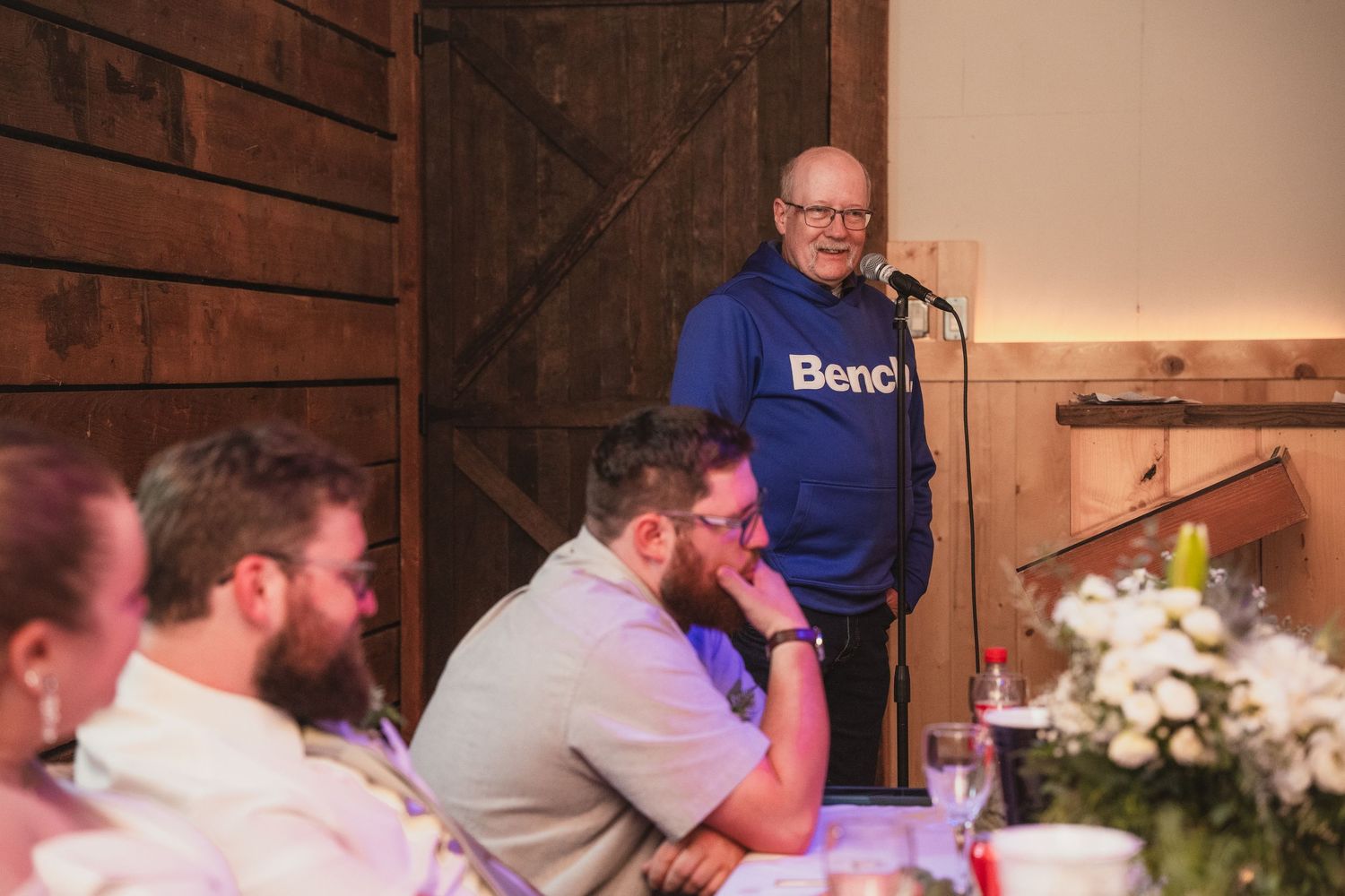 Man in blue Bench hoodie delivers a heartfelt speech at a rustic barn wedding reception.