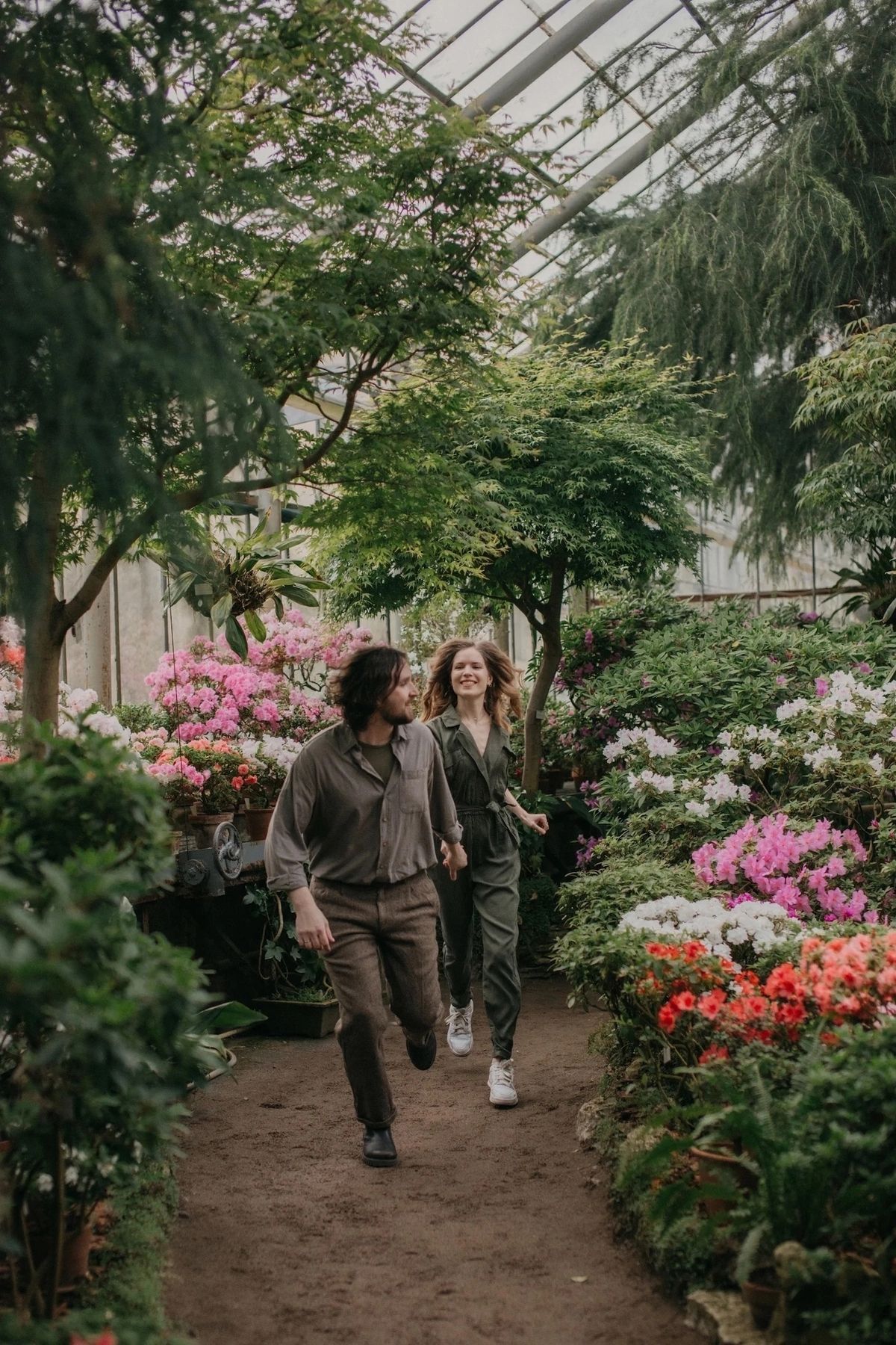 Couple walking hand-in-hand through a lush greenhouse filled with colorful blooming flowers and tall green trees.