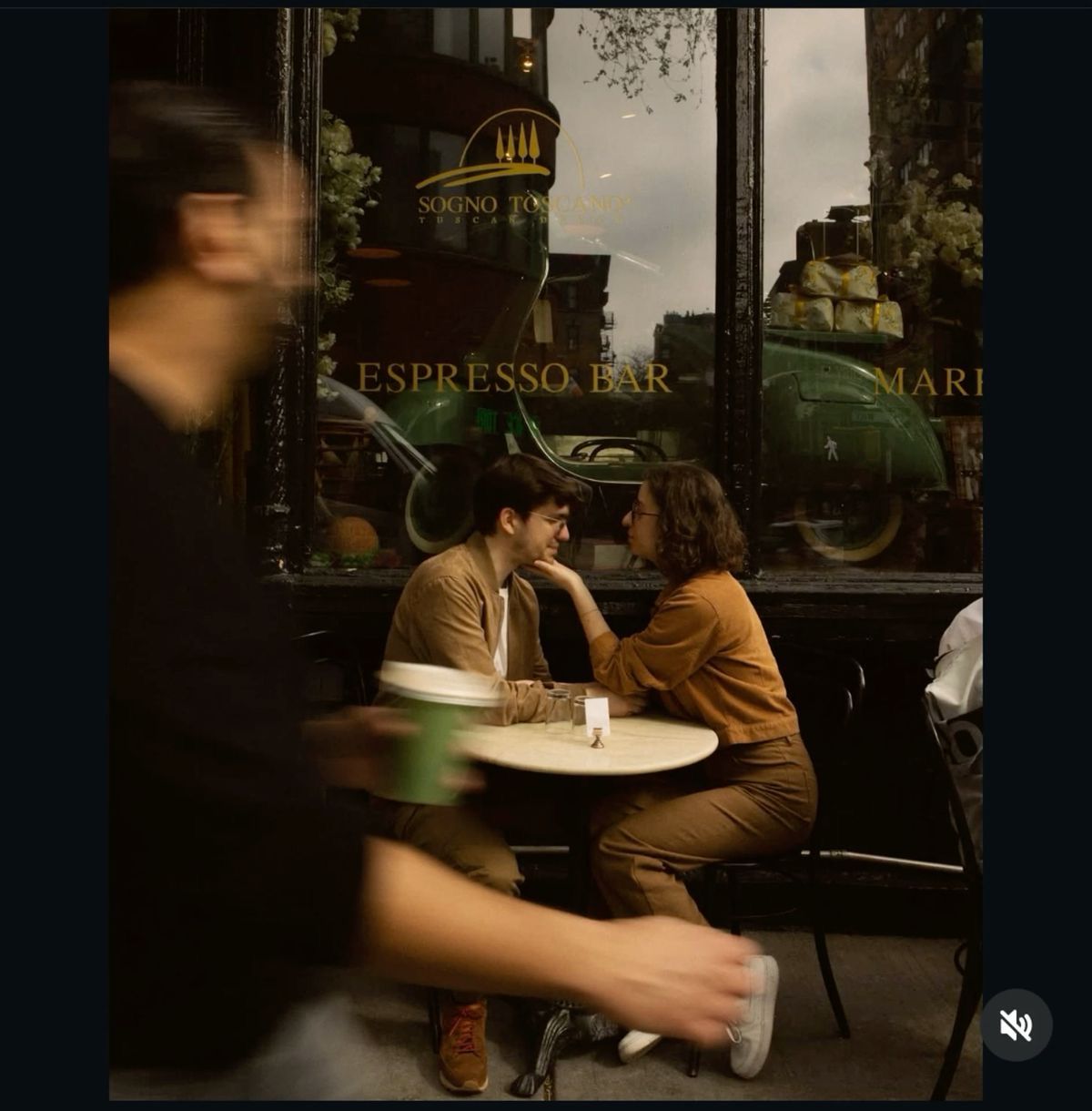 Couple sitting intimately at small table outside espresso bar cafe, captured through window in candid street photography.