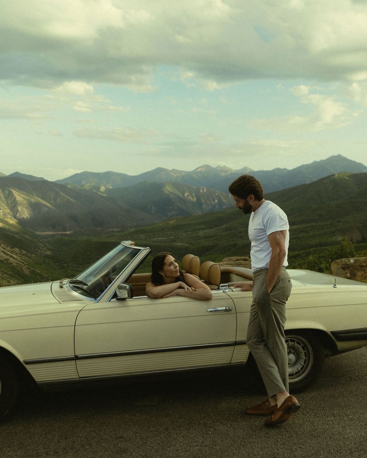 Couple enjoying a scenic mountain view beside a classic white convertible car on a sunny day.