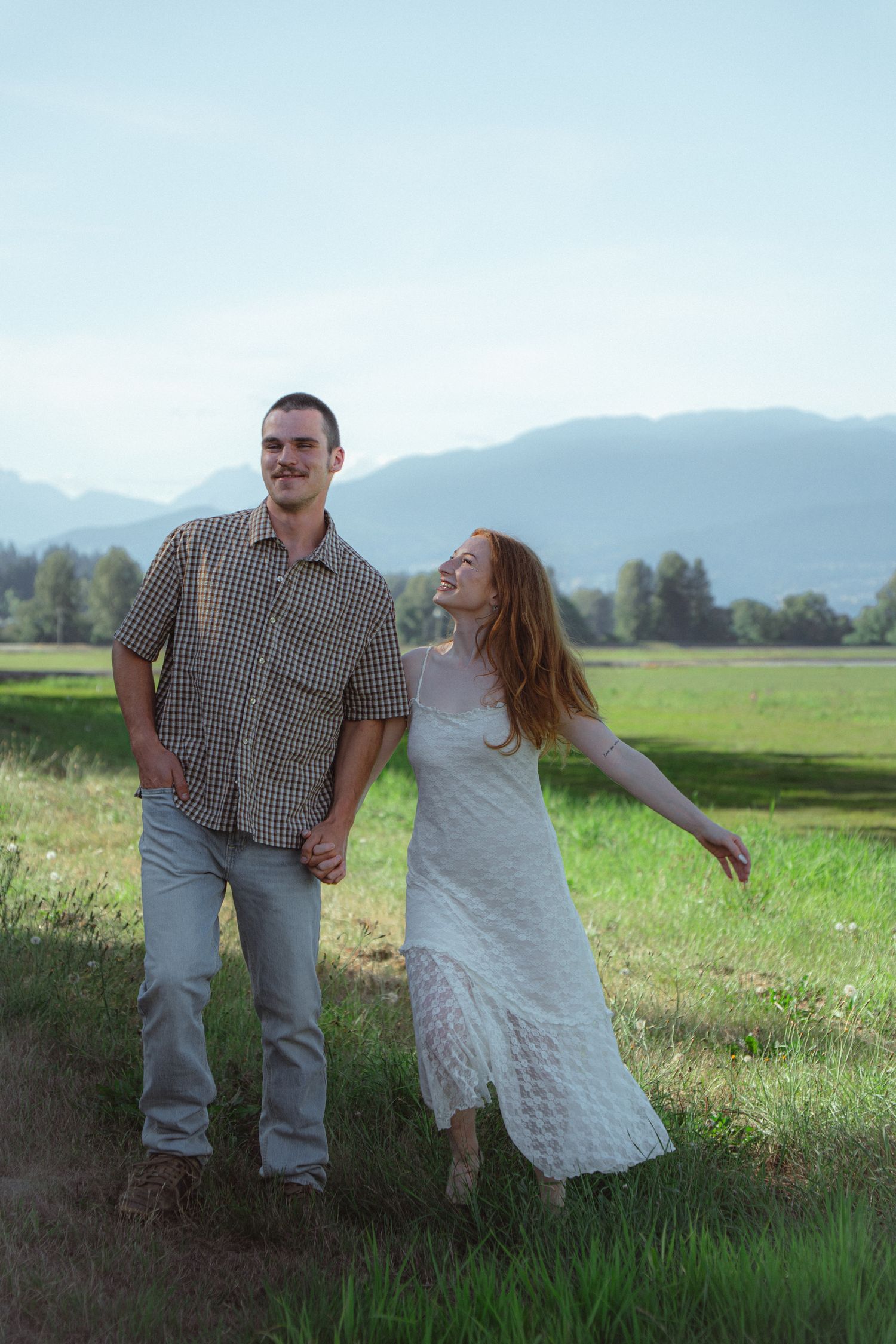 Happy couple holding hands in a green meadow with mountain backdrop, woman in white dress laughing joyfully.