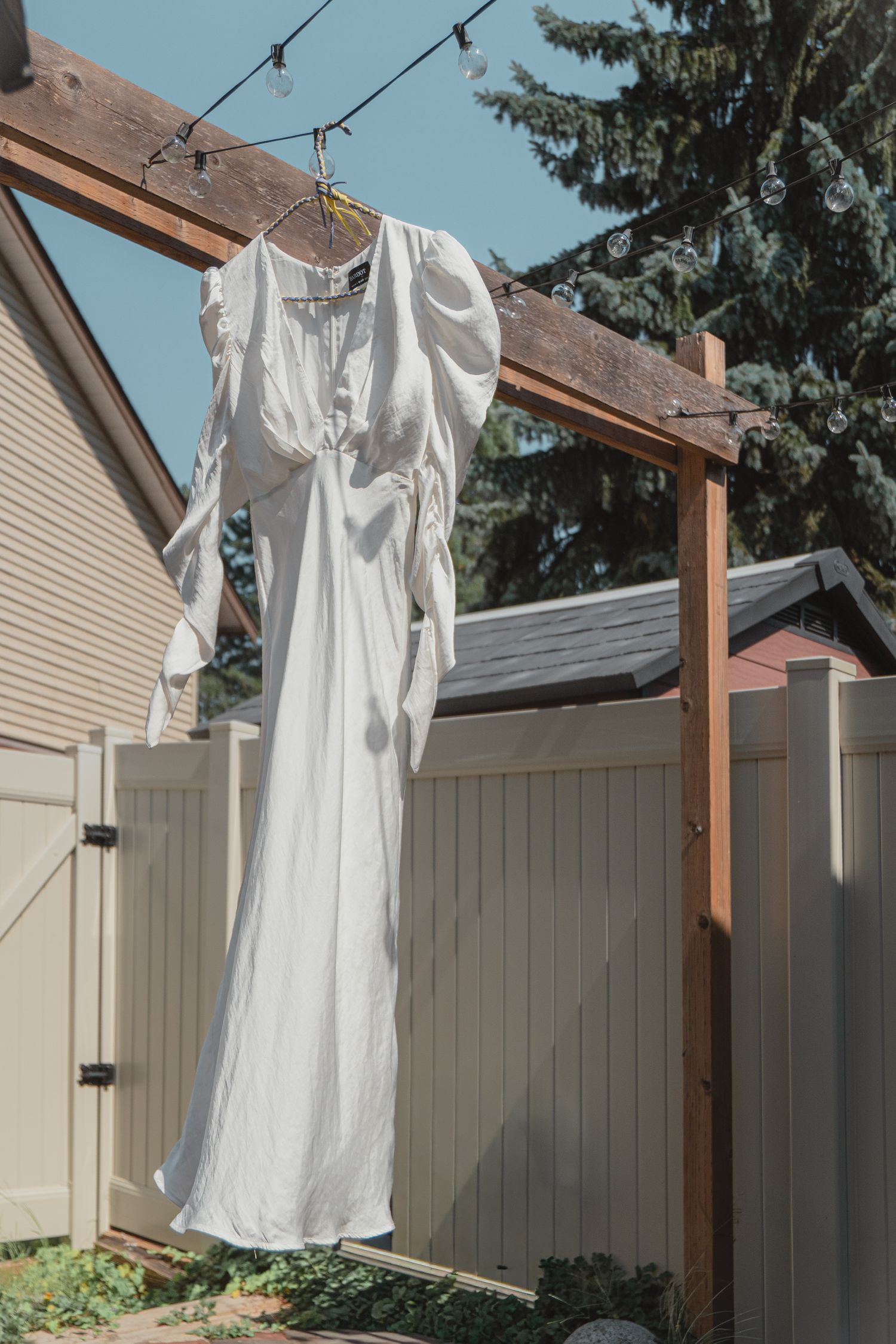 White wedding dress hanging on a wooden pergola outdoors against a blue sky with evergreen trees in the background.