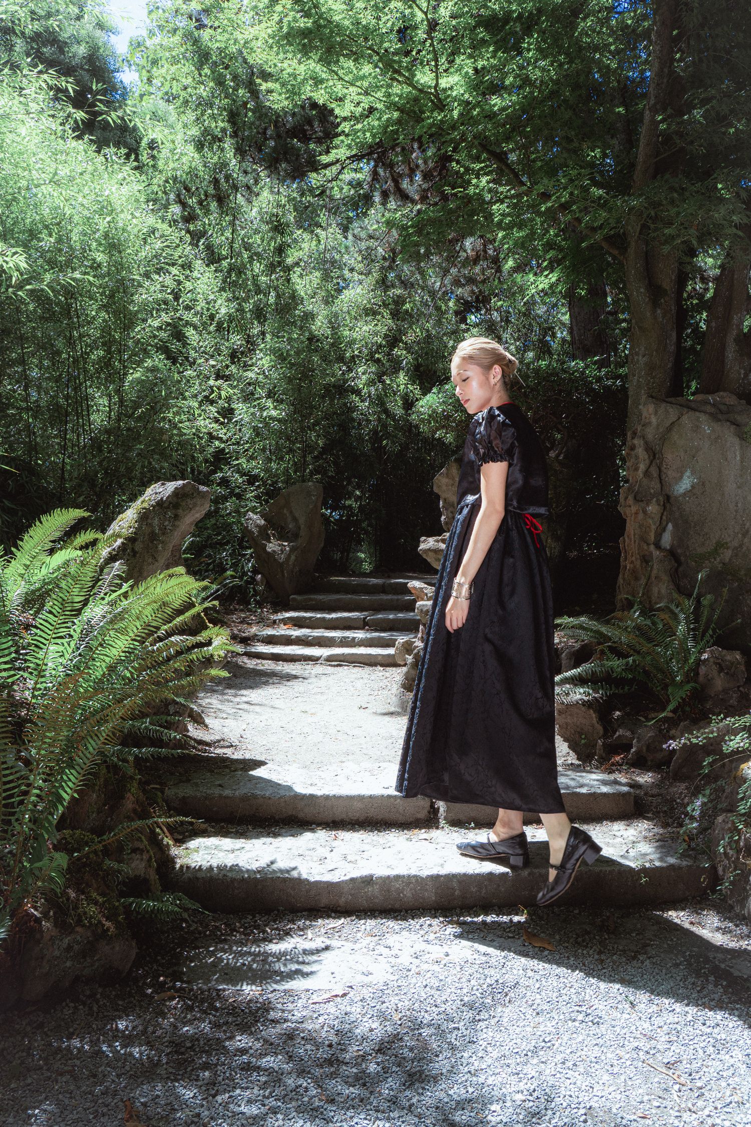 Woman in black midi dress standing on stone garden path surrounded by lush green ferns and trees.