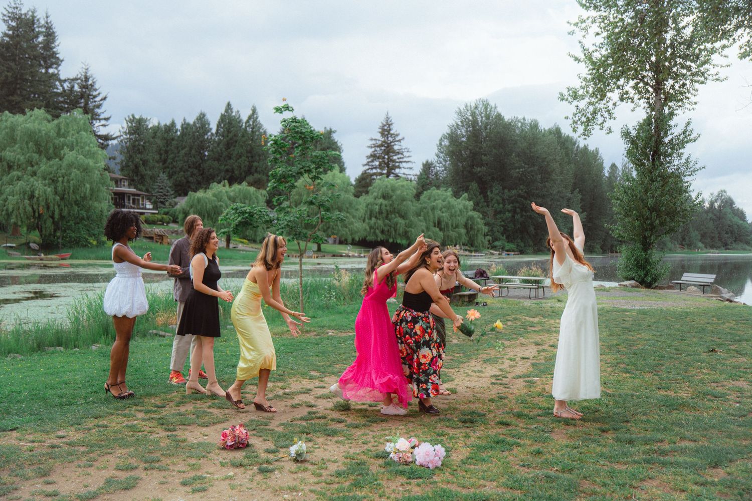 Bridesmaids reaching for bouquet toss on grass with lake and willow trees in background.