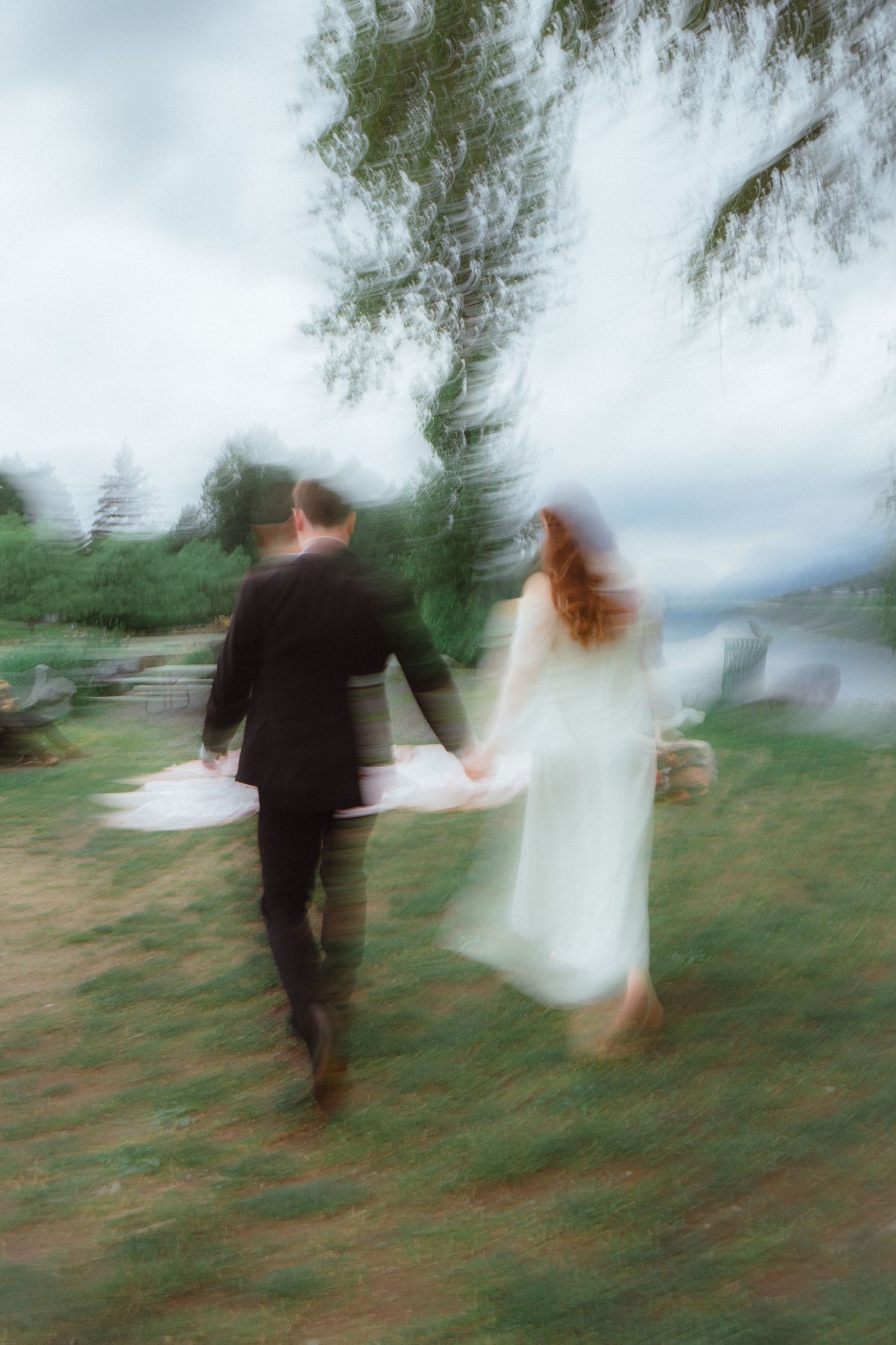 Blurred artistic photo of bride and groom holding hands walking through garden near mountain lake.