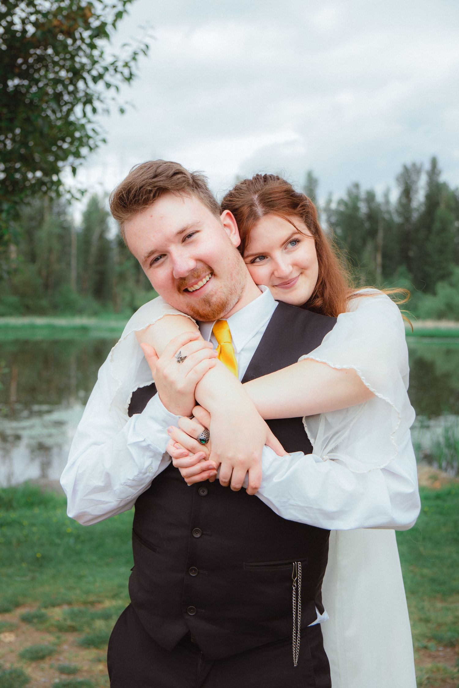 Smiling groom in black vest and yellow tie being hugged from behind by bride near a lake.