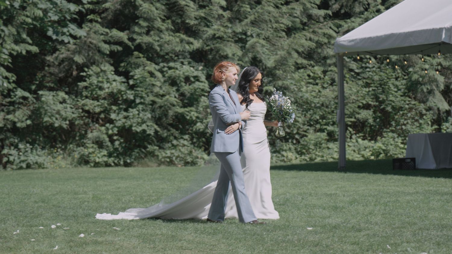Bride in white gown and groom in gray suit walk together on green lawn at outdoor wedding.