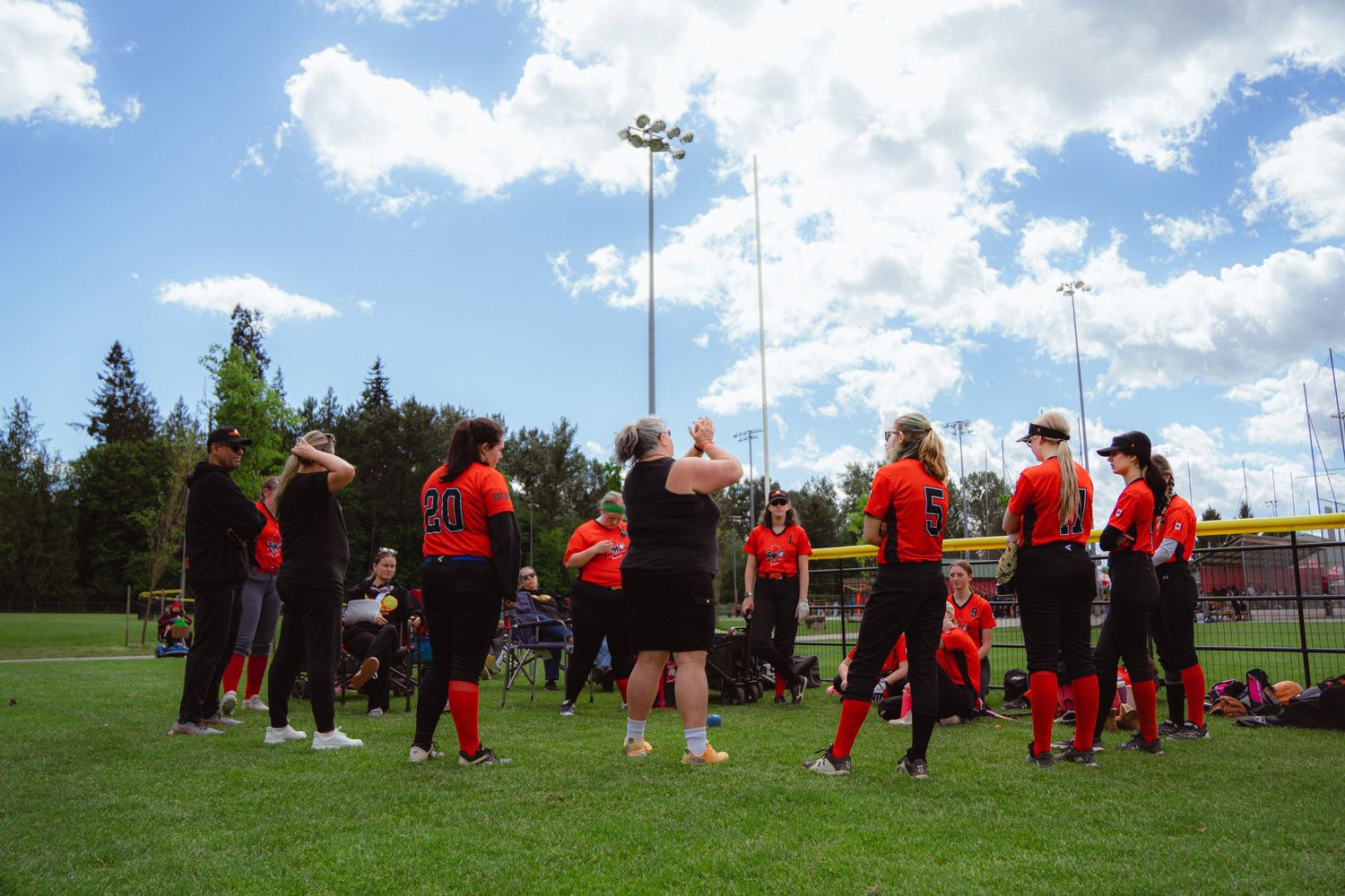 Female softball team in orange and black uniforms gathered together on green field outdoors.