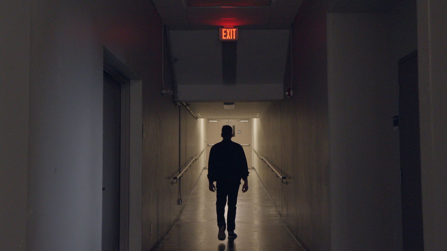 Person walking away down a dark corridor toward a glowing red exit sign on the ceiling.
