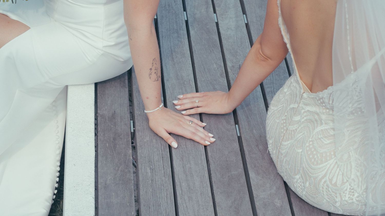 Close-up of two brides in white wedding dresses holding hands on a wooden deck, showing wedding rings.
