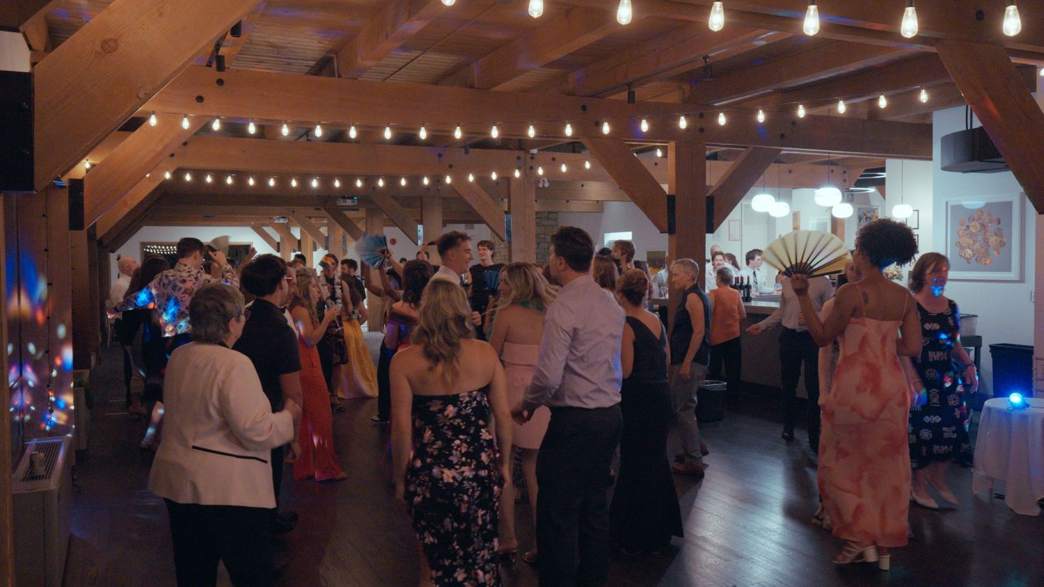 Wedding reception guests mingle and dance under warm string lights in a rustic barn venue.