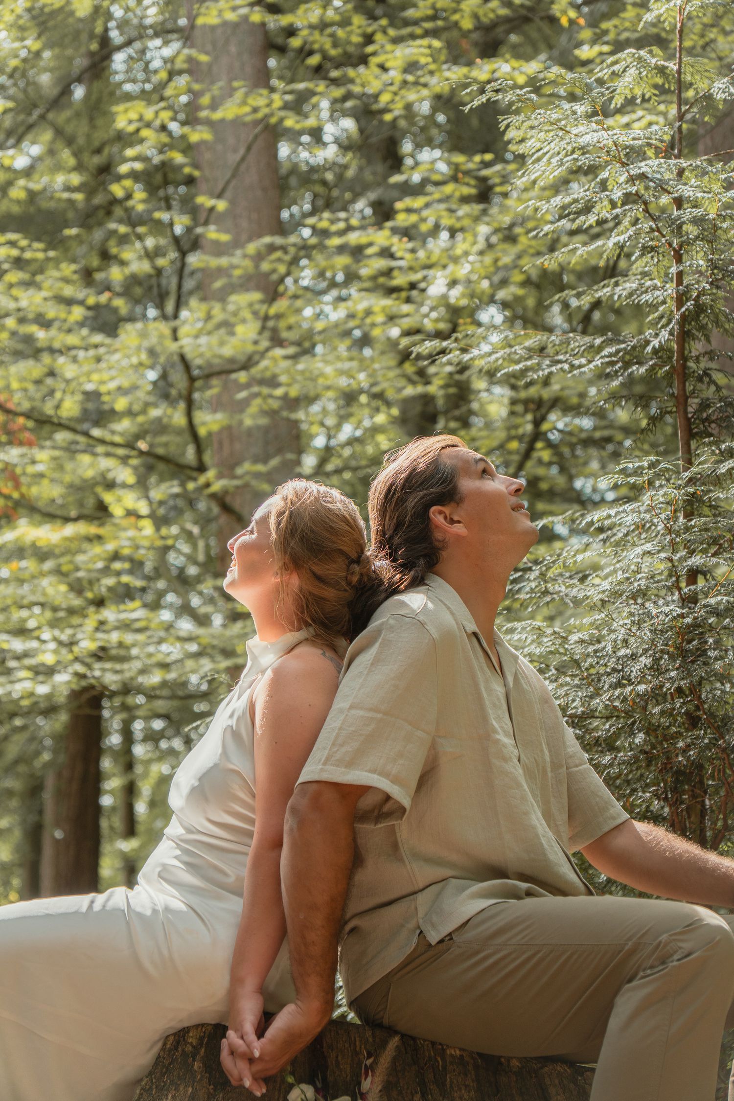 Couple sitting back-to-back on a log, looking up through sunlit green forest trees.