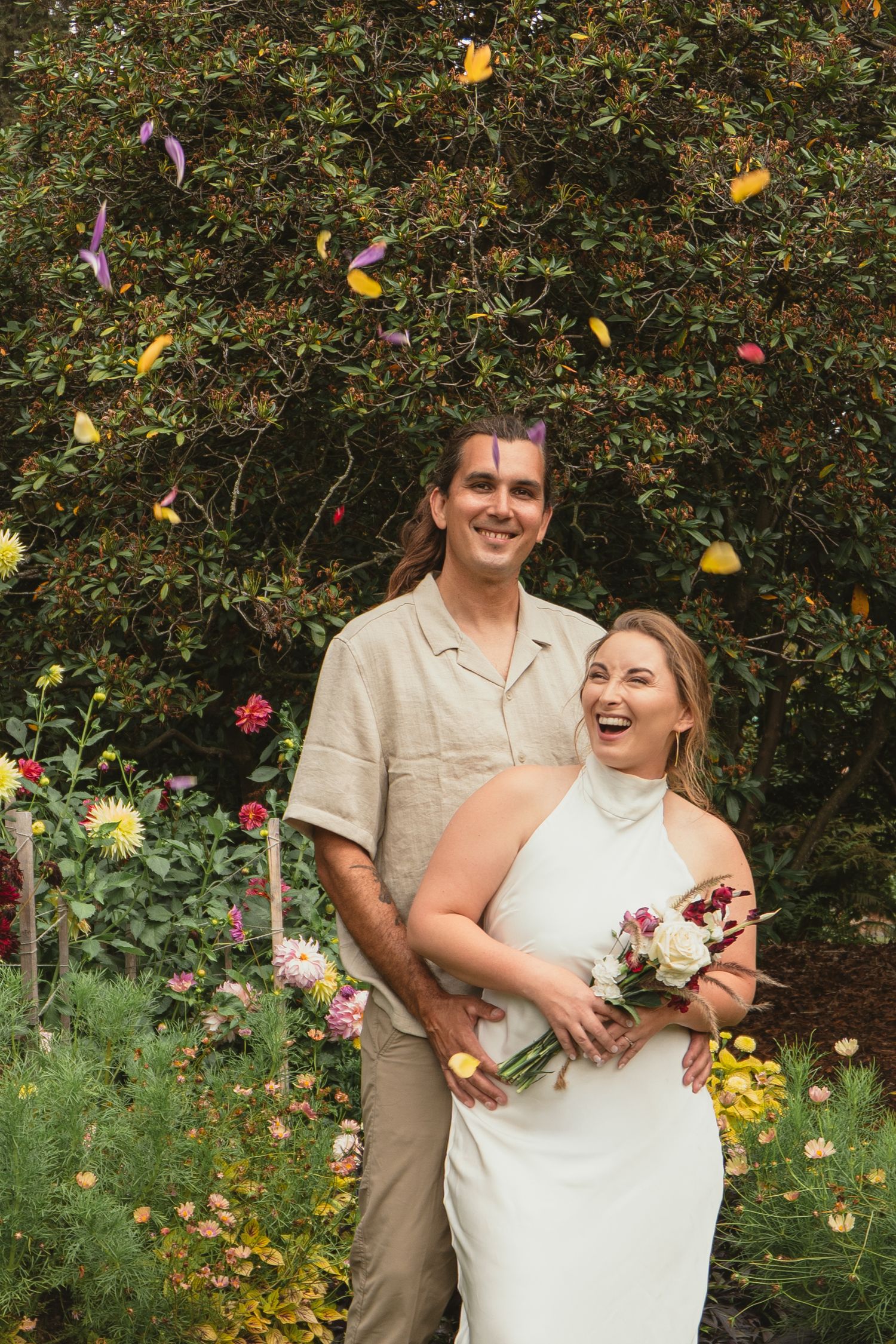 Joyful newlyweds celebrate with colorful confetti in a lush garden, bride holding bouquet in white halter gown.