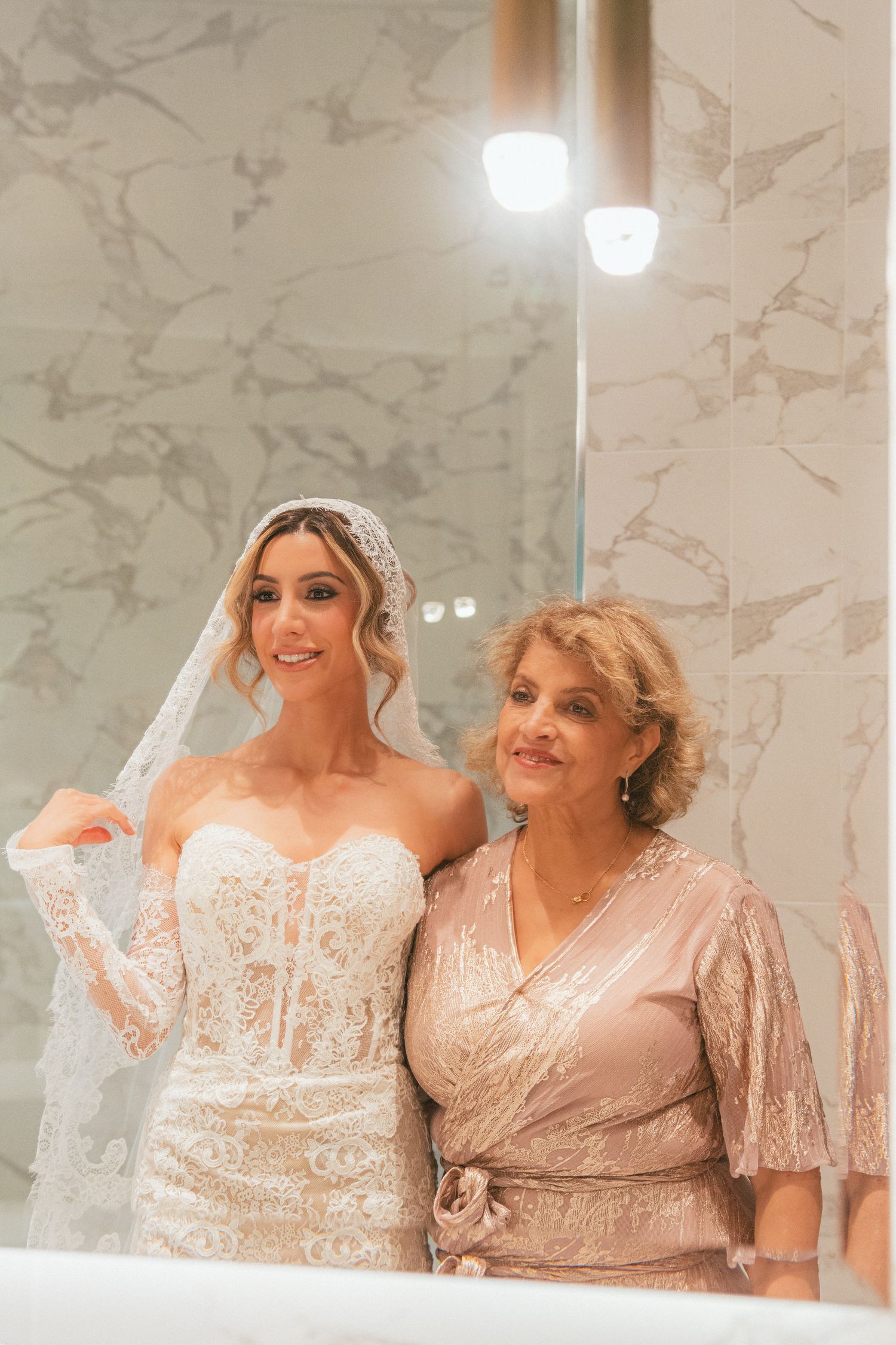 Bride in lace wedding dress and veil poses with older woman in champagne gown in elegant marble bathroom.