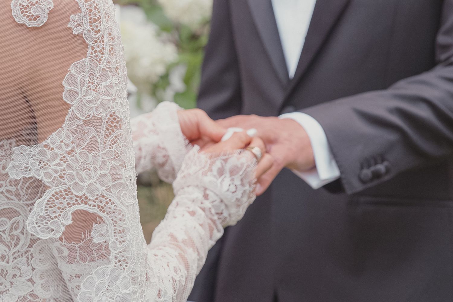 Bride in lace gown and groom in gray suit exchanging wedding rings during outdoor ceremony.