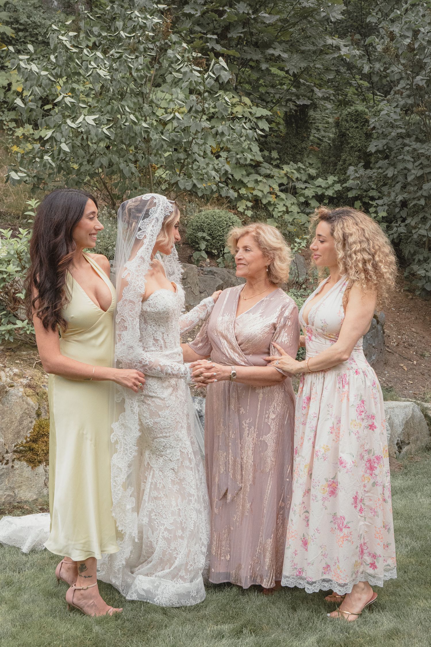 Bride in lace gown and veil stands with three women in pastel dresses in a garden setting.