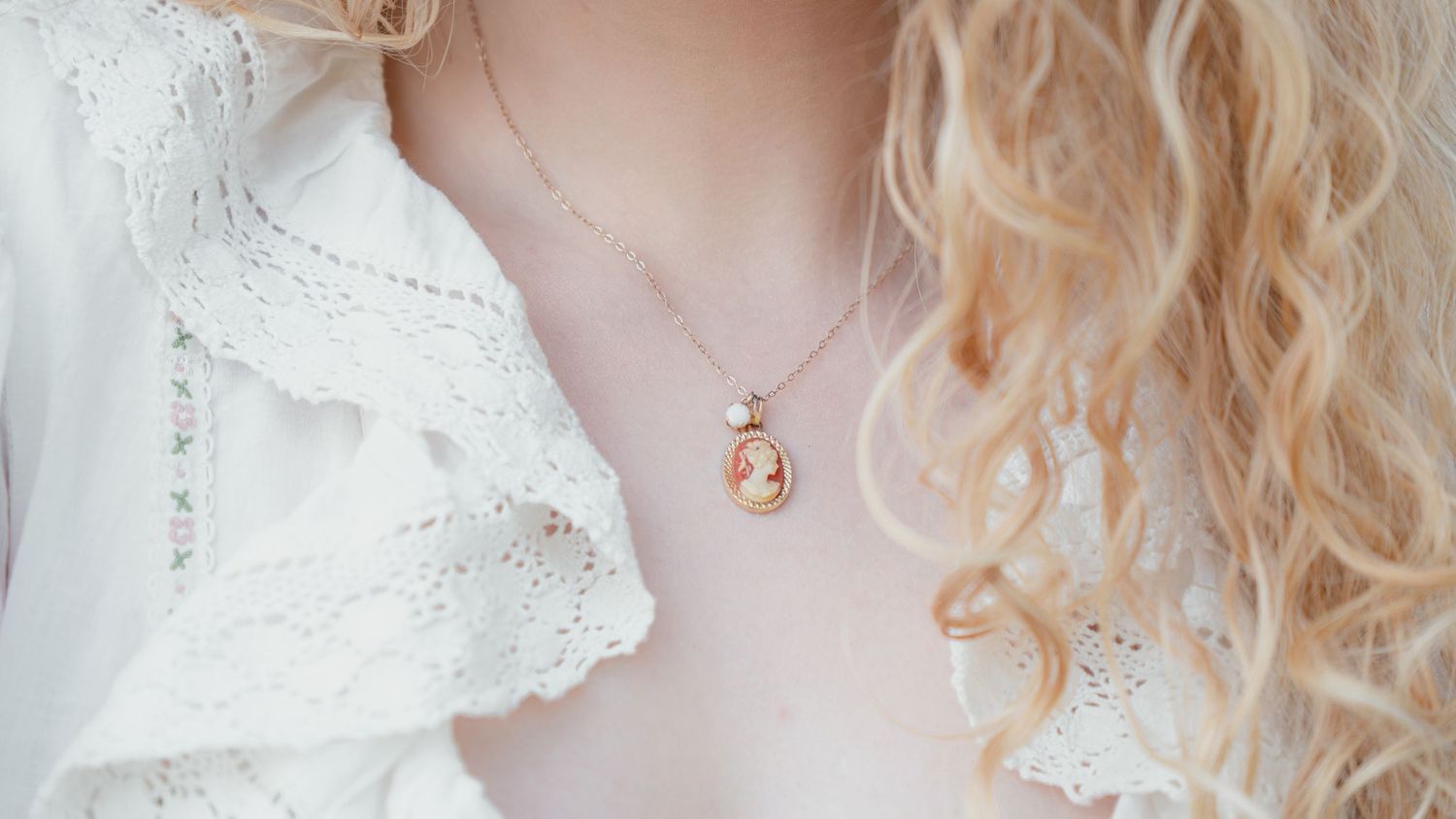 A woman wearing a delicate silver cameo pendant necklace over a white lace blouse with curly blonde hair.