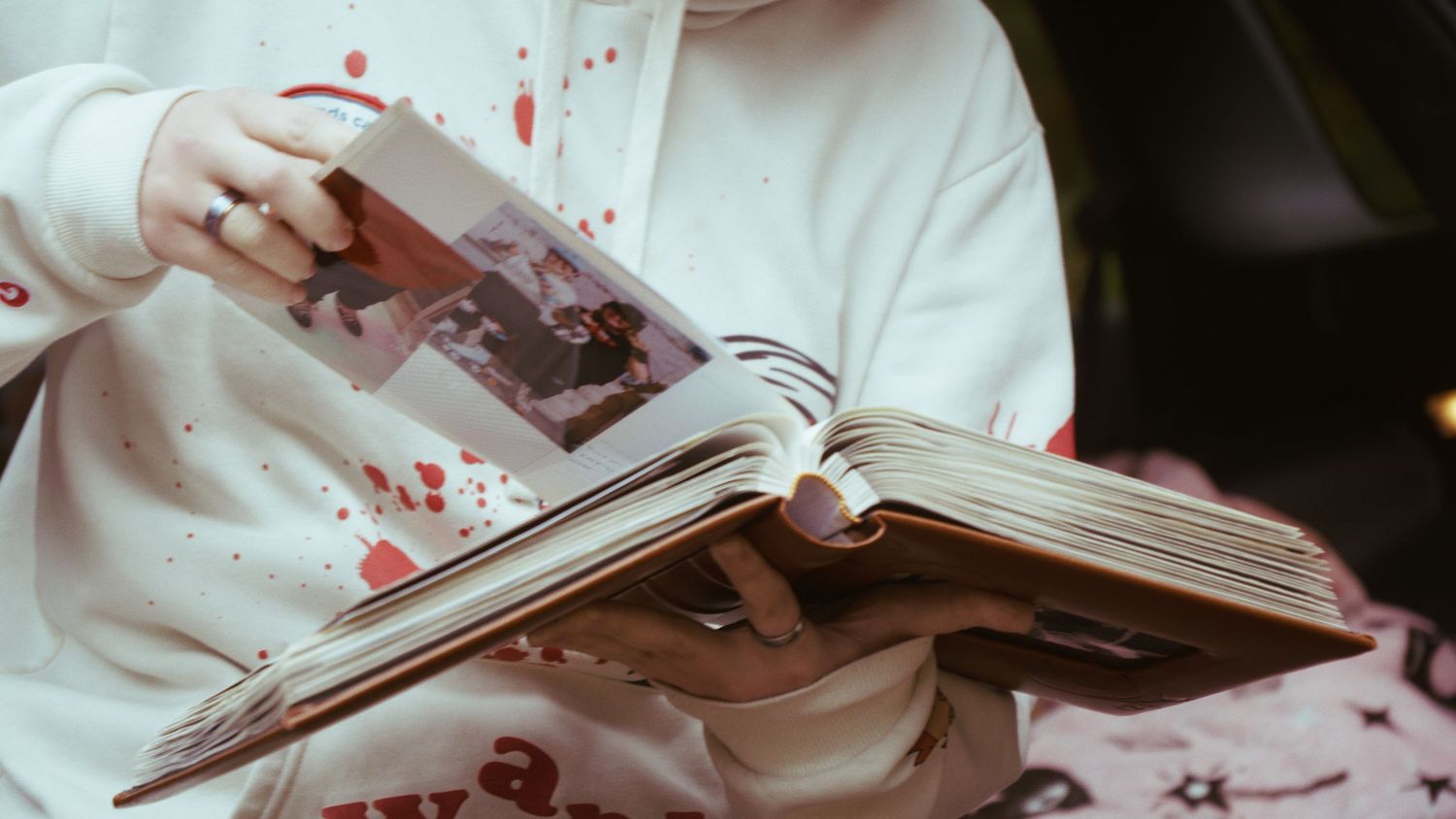 Person in white hoodie with red paint splatter holds open a vintage photo album.