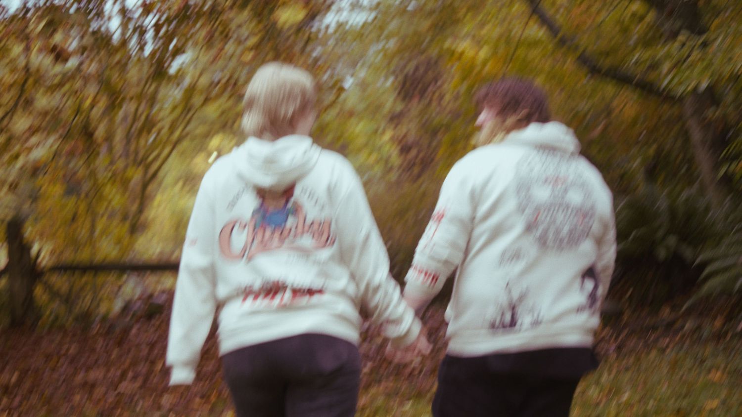 Two people holding hands from behind, wearing matching white hoodies, walk along an autumn forest path.