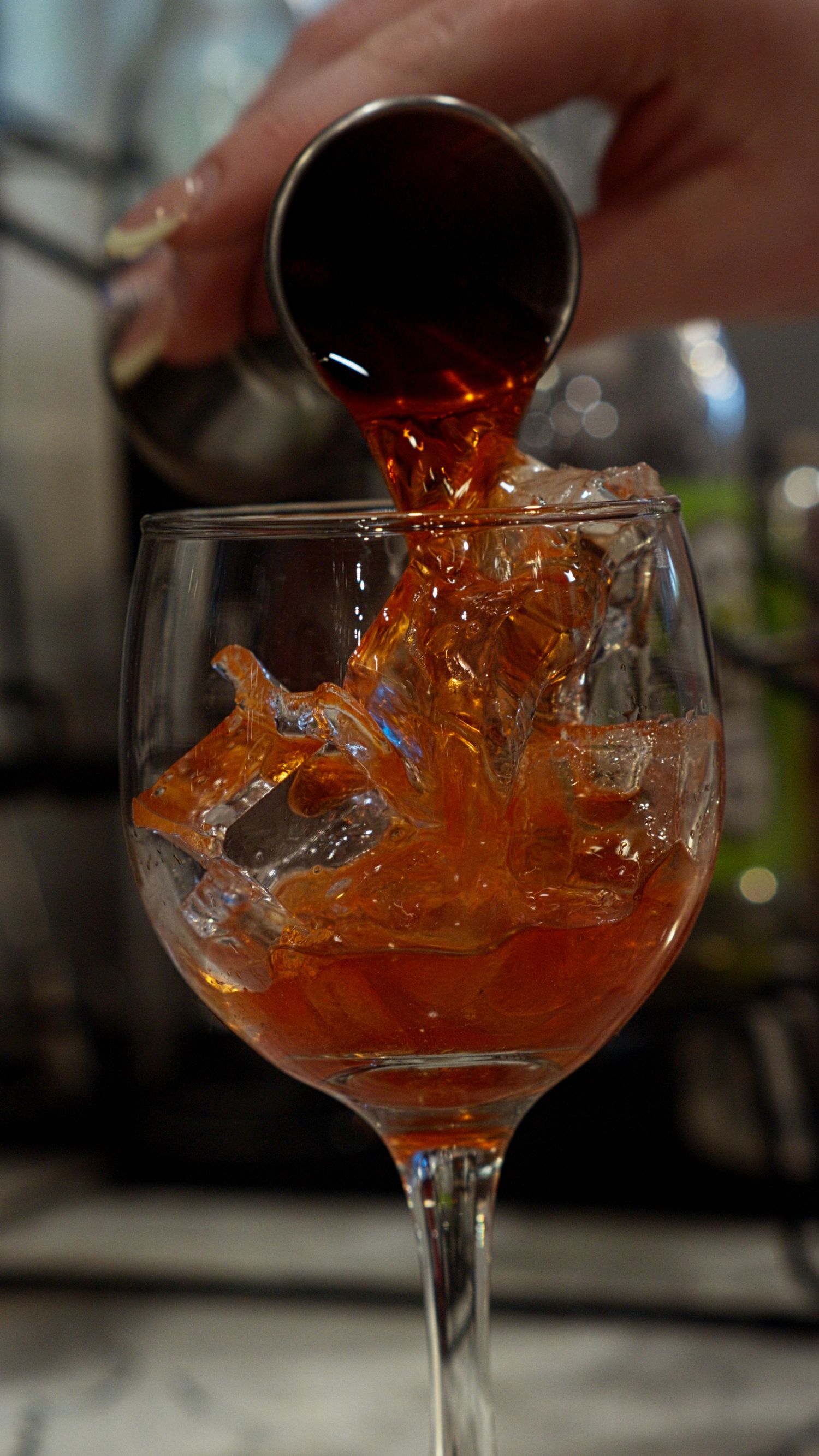 Hand pouring deep amber cocktail over ice into a wine glass with a dramatic splash effect.