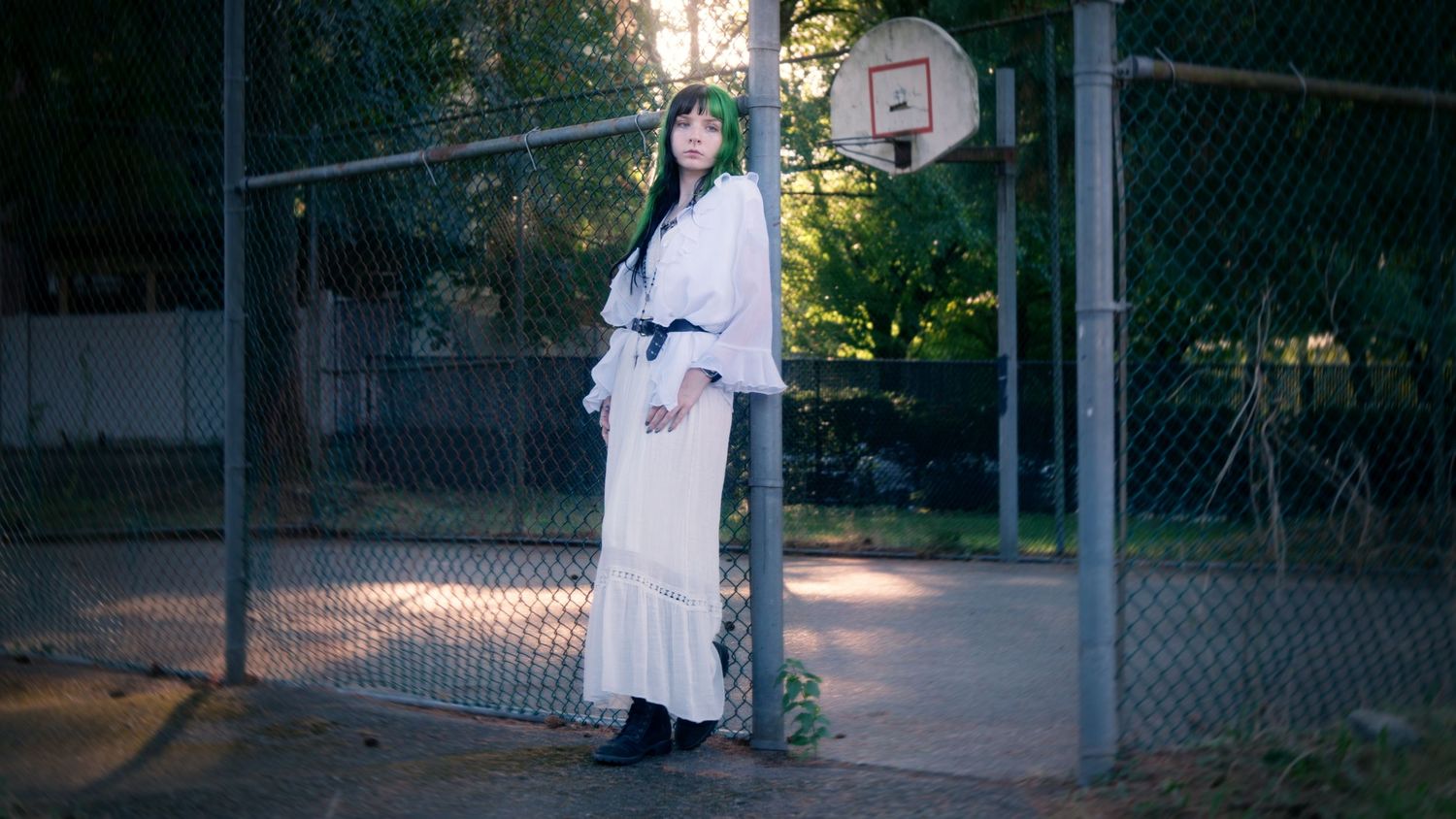 Woman in white dress and black belt poses by chain-link fence near basketball court.