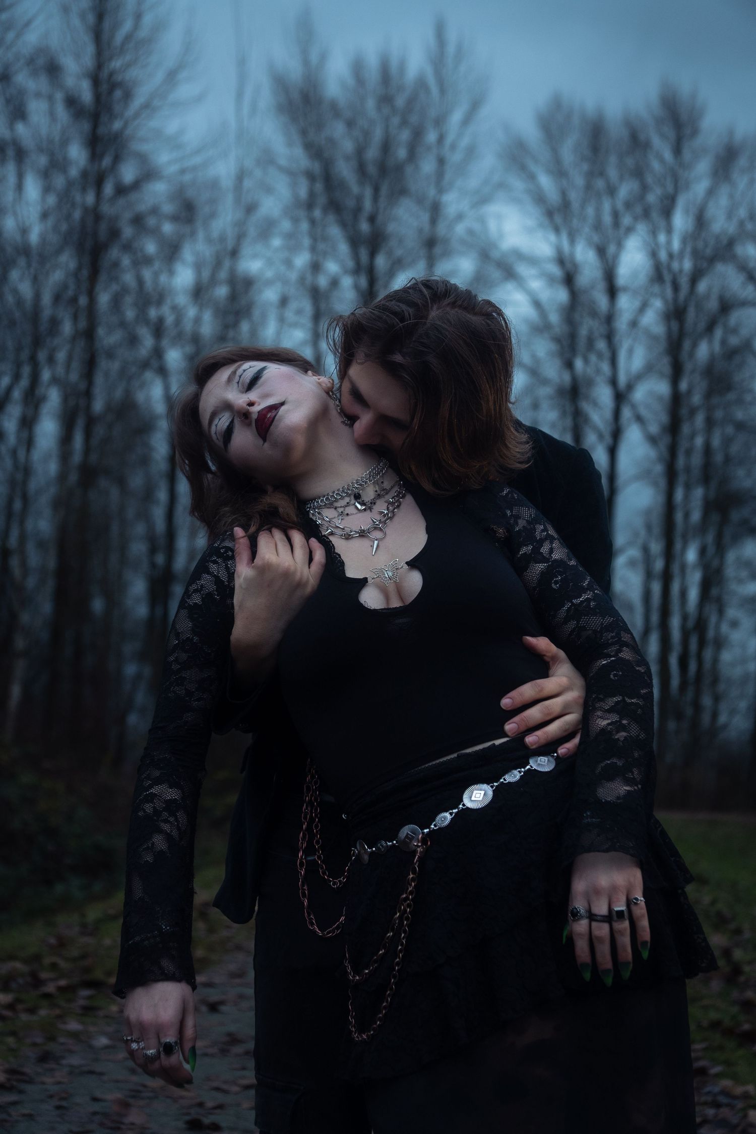Gothic couple in black embrace dramatically on a forest path under a moody cloudy sky.