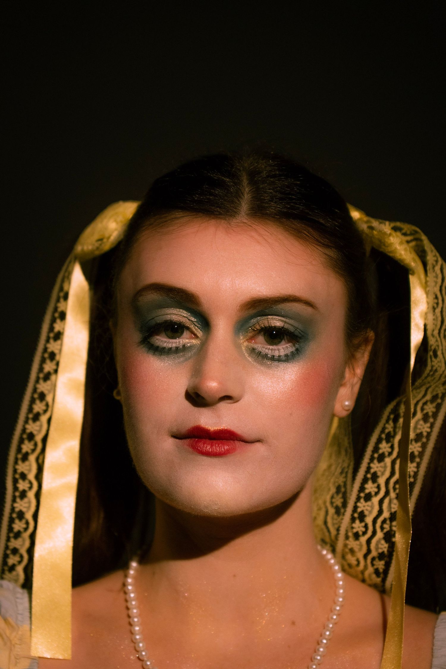 Woman with dramatic stage makeup, blue eyeshadow, red lips, pearl necklace, and gold lace veil ribbons on dark background.