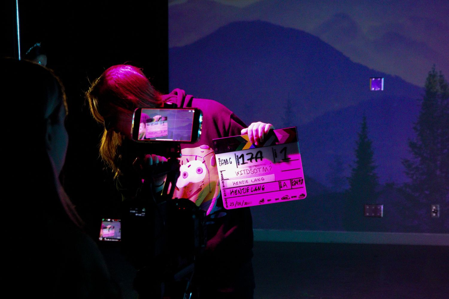 Camera operator filming with a clapperboard in a dark studio with mountain backdrop.