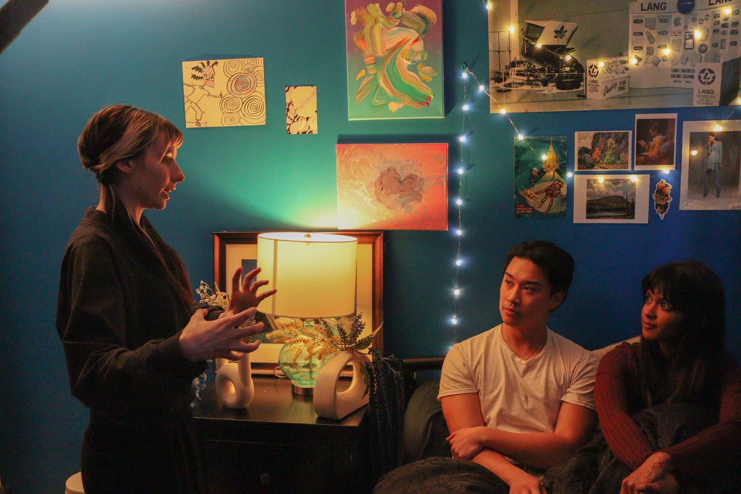 A woman speaks to two seated guests in a cozy room decorated with colorful artwork and string lights.