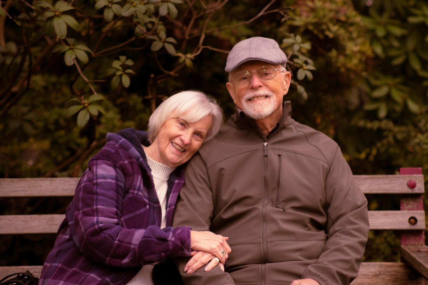 Happy elderly couple sitting on a park bench in fall, smiling and holding hands surrounded by autumn foliage.
