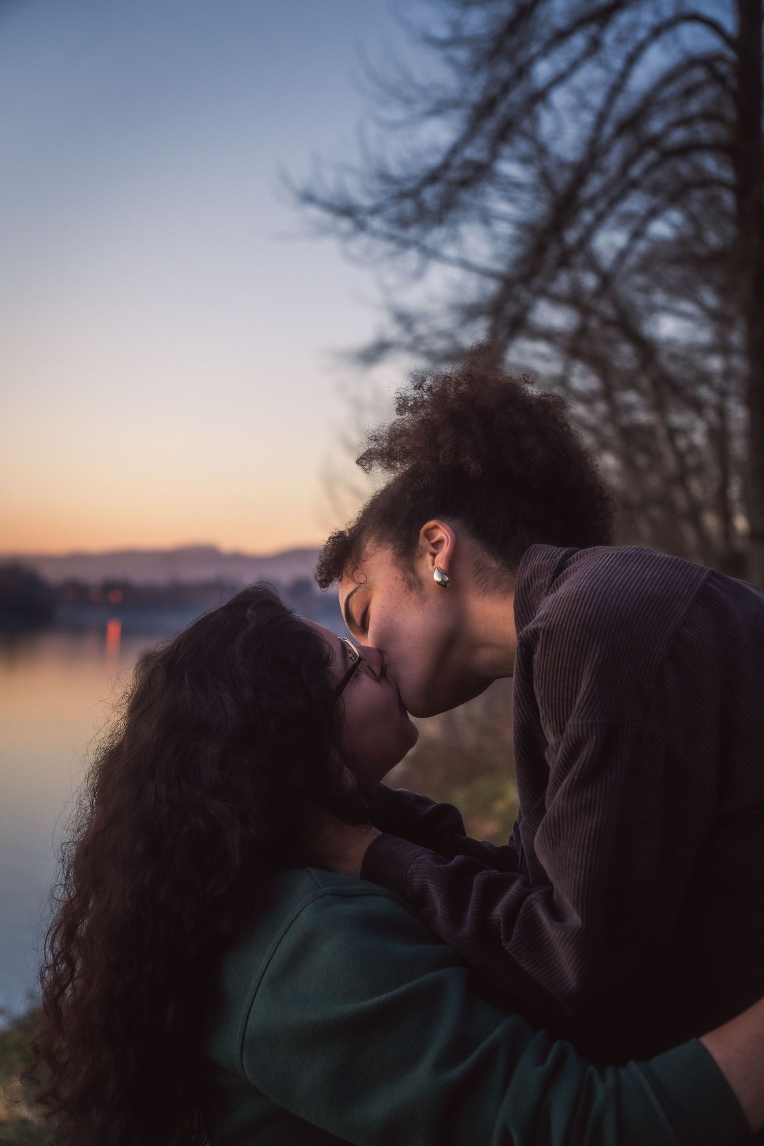 Two women sharing a romantic kiss by a tranquil lake at sunset.
