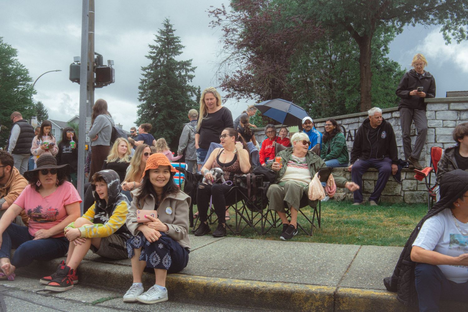 Diverse crowd of spectators sit and stand along a curb watching a community parade on a cloudy day.