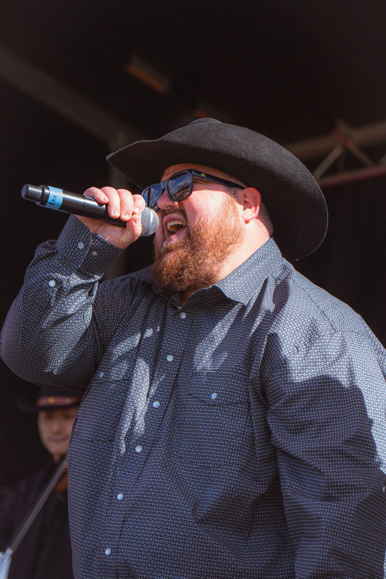 Bearded male country singer in black cowboy hat and sunglasses performs with microphone on stage.