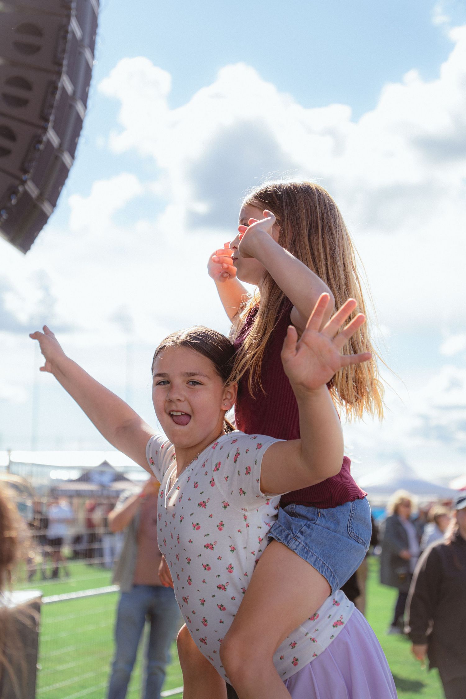 Excited girl carrying friend on shoulders at outdoor music festival under blue sky.