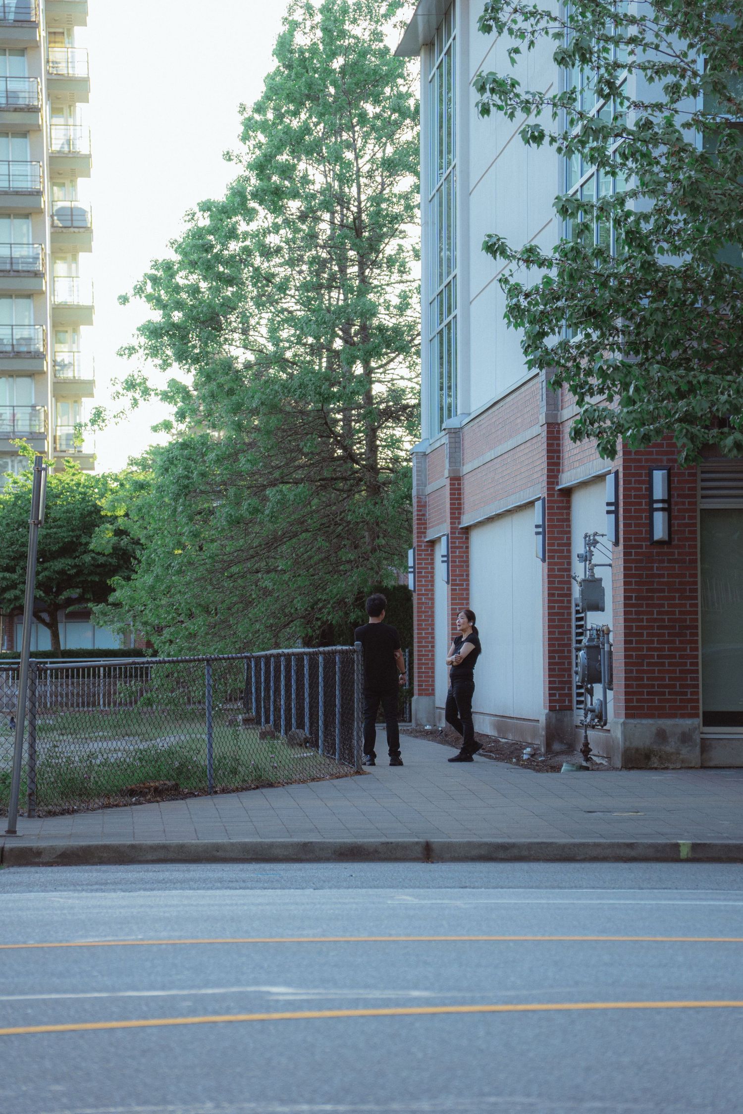 Two people stand on a city sidewalk near brick and concrete buildings with green trees.
