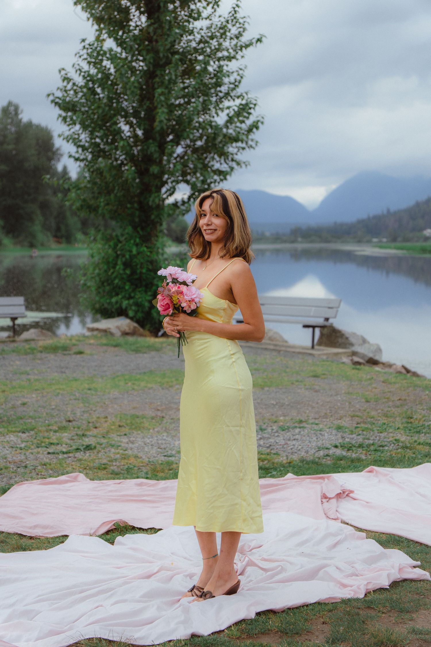 Young woman in yellow midi slip dress holds pink flowers by a peaceful mountain lake.